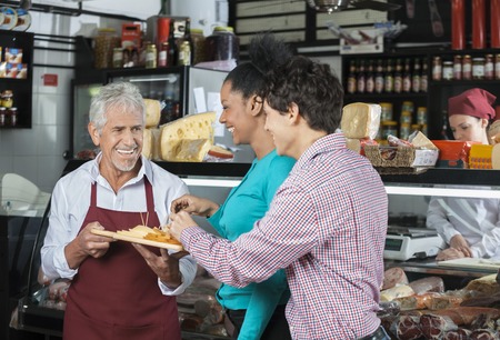 Happy male salesperson offering free samples to customers in cheese shopの写真素材