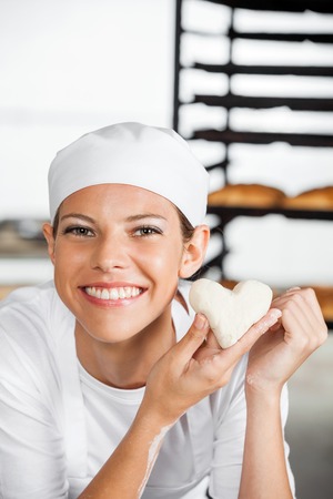Portrait of beautiful baker holding heart shape dough in bakeryの写真素材