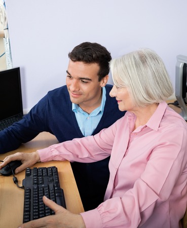 Young male teacher assisting senior woman in using computer at classroomの写真素材