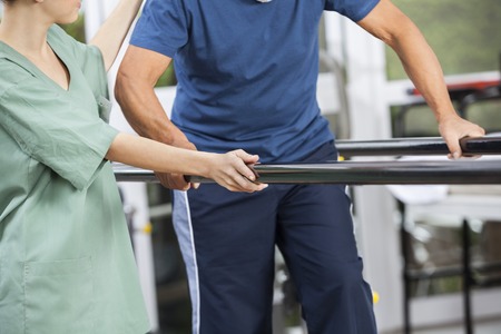 Midsection of female physiotherapist standing by senior man walking between parallel bars in fitness centerの写真素材