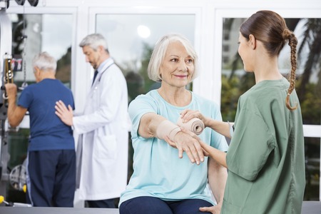 Smiling female patient looking at nurse putting crepe bandage on hand at fitness centerの写真素材