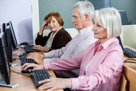 Portrait of smiling senior woman sitting in computer class with classmates studying at deskの写真素材