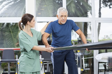 Female physiotherapist motivating senior patient to walk between parallel bars in rehabilitation centerの写真素材