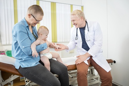 Happy baby touching doctor's hand while sitting on mother's lap in hospitalの写真素材