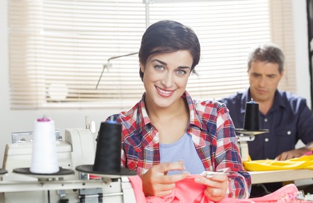 Portrait of confident female tailor sitting at workbench with colleague working in background at...