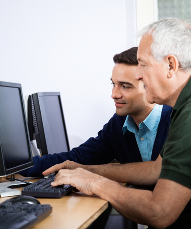 Young male teacher assisting senior man in using computer at classroomの写真素材
