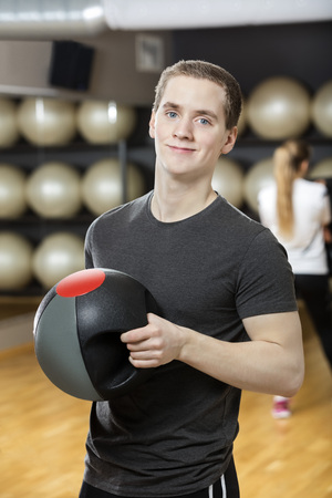 Portrait of young man smiling while holding medicine ball at gymnasiumの写真素材