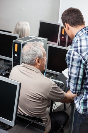 Male tutor assisting senior man in using computer at classroomの写真素材