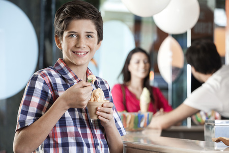 Portrait of smiling preteen boy having ice cream while mother and waiter standing at counter in parlorの写真素材