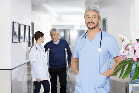 Portrait of confident doctor standing with hands in pockets while colleague and senior patient communicating in background at rehab centerの写真素材