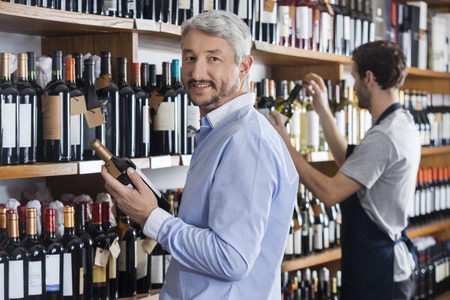 Portrait of mature customer holding wine bottle while salesman working in shopの写真素材