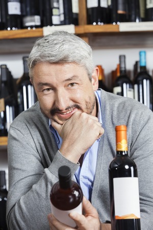 Portrait of happy mature man with hand on chin holding wine bottle in shopの写真素材