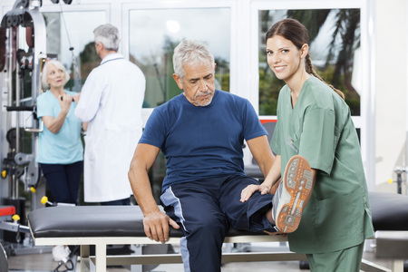 Smiling female nurse assisting senior man in leg exercise at rehab fitness centerの写真素材