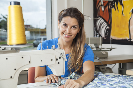 Portrait of smiling female tailor stitching fabric at workbench in sewing factory