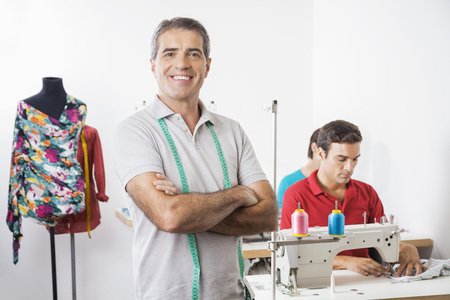 Portrait of confident male tailor standing arms crossed at factory with employees working in backgroundの写真素材