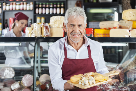 Portrait of smiling salesman holding various cheese on cutting board with colleague in background at...