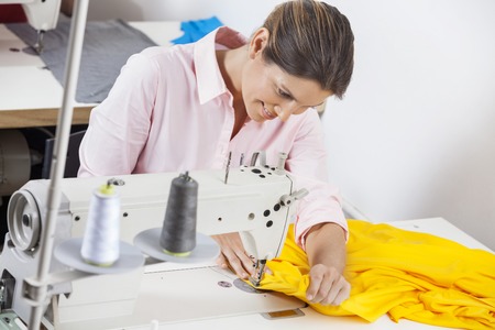 Smiling female tailor stitching yellow fabric at workbench in factoryの写真素材