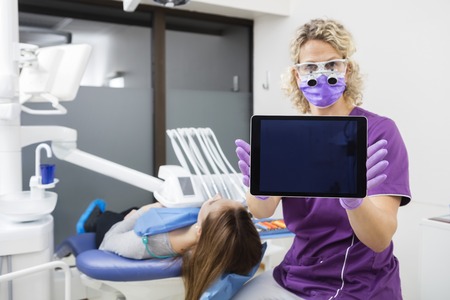 Portrait of confident female dentist showing digital tablet with blank screen by patient in clinicの写真素材