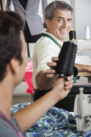 Happy mature tailor giving thread spool to male colleague in sewing factory