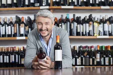 Portrait of happy mature man holding wine bottle while leaning on table in shopの写真素材