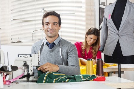 Portrait of smiling tailor sitting at workbench in sewing factory with colleague working in backgroundの写真素材
