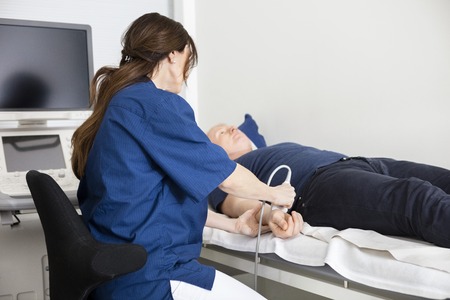 Mature female doctor using ultrasound probe on male's hand in hospitalの写真素材