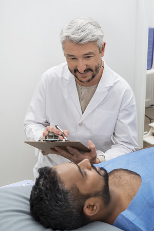 Doctor Writing On Clipboard While Looking At Patient On Bedの写真素材