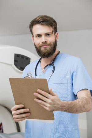 Confident Young Male Doctor With Clipboard In Examination Roomの写真素材