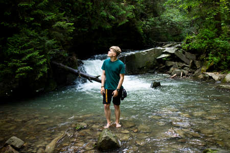 Young guy stands by the river against the background of a waterfallの写真素材