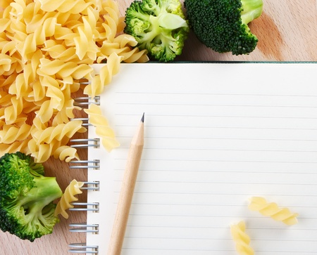 Directly above close-up view of a table with a pasta, broccoli and a notebook on cutting board