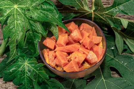 Bowl with pieces of papaya. Tropical fruit. Edible fruit with digestive properties. Green leaves. Wood background. Fruit for juices. Natural food and detox.の写真素材