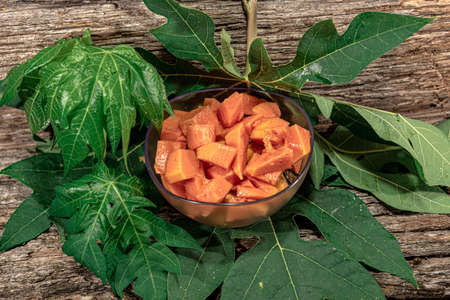 Bowl with pieces of papaya. Tropical fruit. Edible fruit with digestive properties. Green leaves. Wood background. Fruit for juices. Natural food and detox.の写真素材