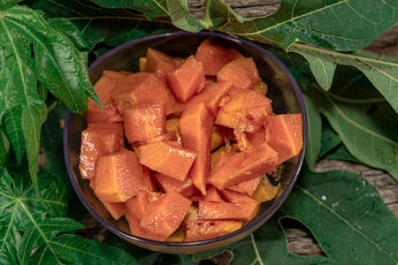 Bowl with pieces of papaya. Tropical fruit. Edible fruit with digestive properties. Green leaves. Wood background. Fruit for juices. Natural food and detox.の写真素材