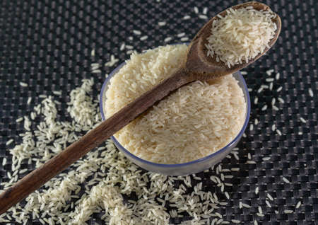 Peeled and processed grain rice on a dark background. Rice cereal and cooking ornaments. Semi-processed grains in a plastic pot and wooden spoons. Foods. Brazilian cuisine.の写真素材