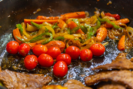 steak with onions, tomatoes and peppers being prepared in a frying pan on a gas stoveの写真素材