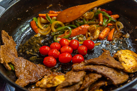steak with onions, tomatoes and peppers being prepared in a frying pan on a gas stoveの写真素材