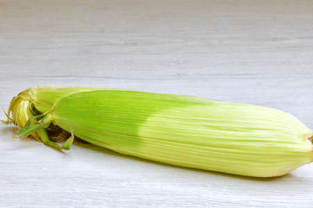 isolated fresh cob of corn in the skin on a light wooden surfaceの写真素材