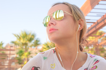 A beautiful young tanned girl listening music with headphones in sunglasses under palms shadow.の写真素材