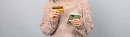 Panorama crop photo of woman holding a credit card and phone in a studio over grey background.