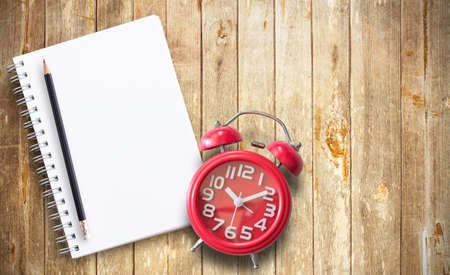 Red alarm clock and note book on brown wooden plank background. Top view with copy space