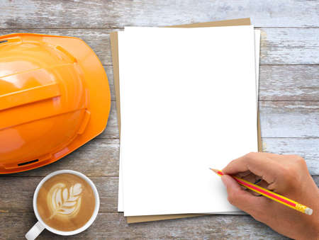 Office desk, Hand holding pencil,safety helmet,blank white and brown paper and coffee on wood table...