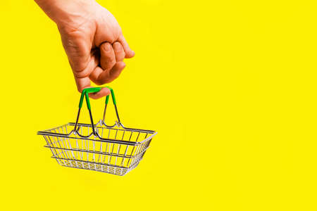 Hand holds shopping basket with its little finger, on yellow background. Concept of a trip to the supermarket.の写真素材