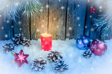 Christmas ornament, red burning candle and toys in the snow on a wooden background and tree branchesの写真素材