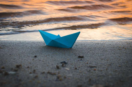 Paper blue ship is thrown on sandy shore of lake at sunset.Beach holiday.の写真素材