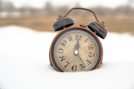 Old retro alarm clock in the snow in winter on the background of vegetationの写真素材