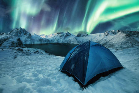 Blue tent camping on snowy hill with Aurora Borealis dancing on mountain range in Senja Island, Norwayの写真素材