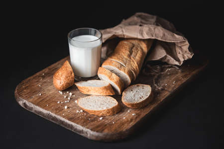 Healthy food. Long loaf of rural bread with two cut-off pieces lie on a wooden chopping board and a glass of fresh milk. Dark background. Horizontal shotの写真素材