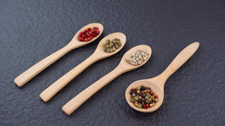 Focus on foreground of various colors peppercorns in wooden spoon on black stone slate backgroundの写真素材