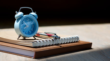 Selective focus at little blue alarm clock with ballpoint pen and kraft paper books with sunlight and shadow on wooden table surface, hobby and relaxing time conceptの写真素材