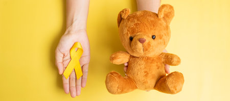 hand holding Yellow Ribbon and Bear doll on yellow background for supporting kid living and illness. September Childhood Cancer Awareness month and World cancer day conceptの写真素材